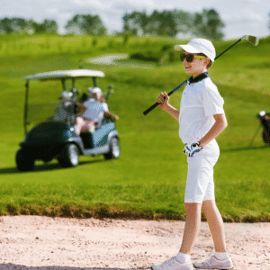 A male child looking happy standing in a golf sand bunker while holding a golf club balanced on his right shoulder with a left hand on his hip. The day is bright and there is a golf buggy in the background on the grass.