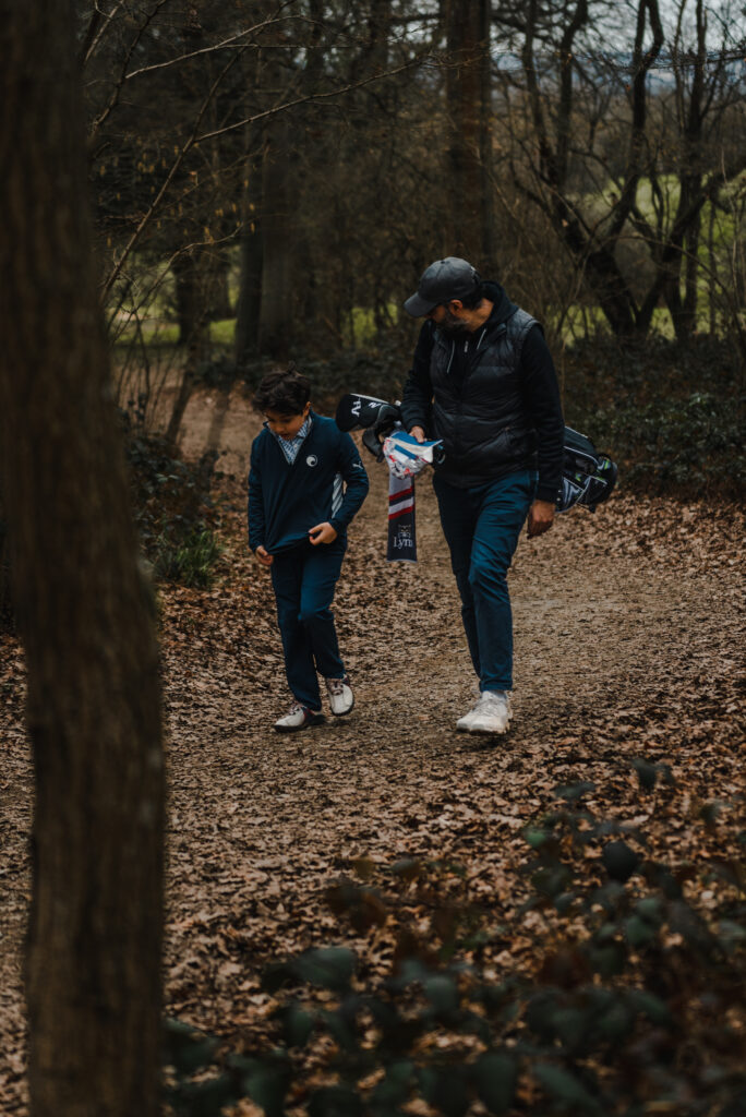 A young junior golfer walks along a wooden path with his dad who is carrying his golf bag.