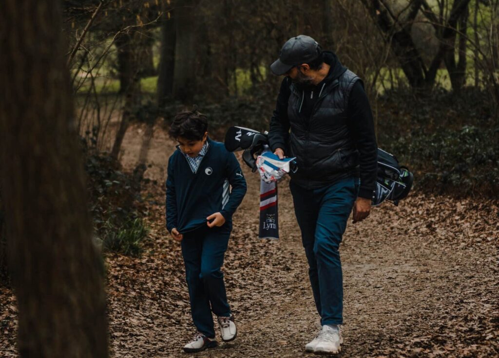 A father and a son walking together through some trees on a golf course, with the father carrying his sons golf clubs.