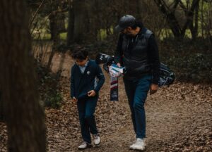 A father and a son walking together through some trees on a golf course, with the father carrying his sons golf clubs.